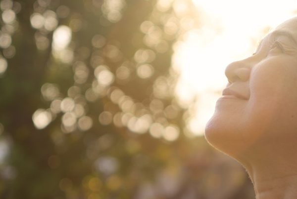 Mulher de meia-idade de olhos fechados e braços abertos, a respirar fundo e a sorrir ao sol, num parque