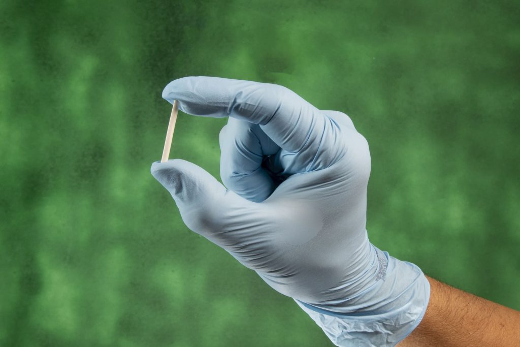 Texto alternativo: "Mão com luva médica azul a segurar um pequeno implante subcutâneo em bastonete, conhecido como chip da beleza, sobre fundo verde.