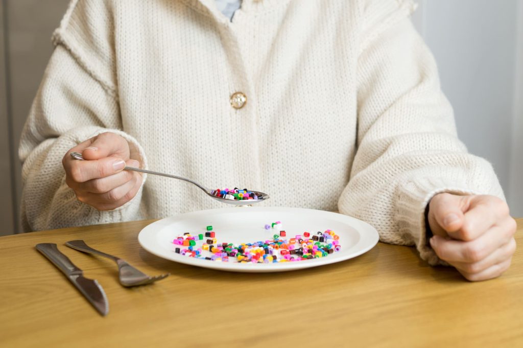 Pessoa à mesa com um prato de comida, a levar à boca uma colher cheia de missangas de plástico coloridas em vez de alimentos, simbolizando a ingestão de microplásticos e disruptores endócrinos