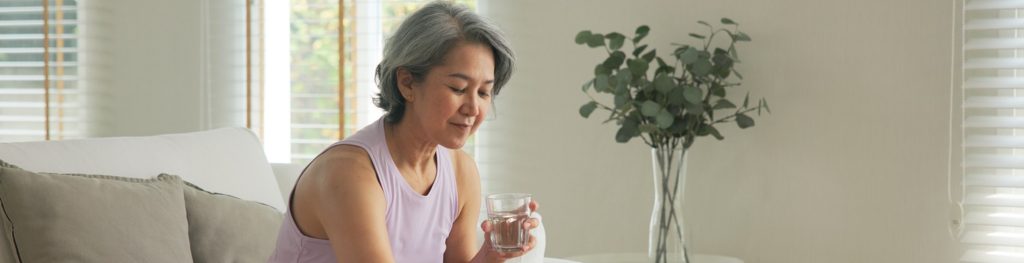 Mulher madura sentada no sofá, com um copo de água na mão, a preparar-se para tomar medicamentos colocados sobre a mesa.