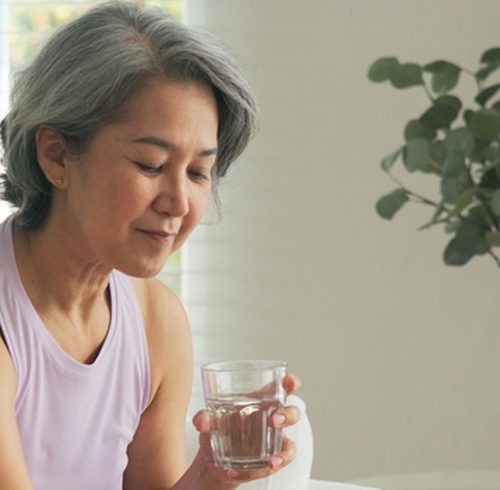 Mulher madura sentada no sofá, com um copo de água na mão, a preparar-se para tomar medicamentos colocados sobre a mesa.
