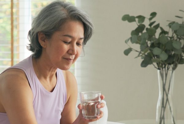 Mulher madura sentada no sofá, com um copo de água na mão, a preparar-se para tomar medicamentos colocados sobre a mesa.